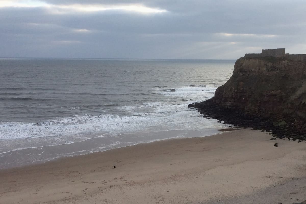 The quiet sands of King Edward’s Bay in Tynemouth on a calm winter day, with a single walker at the water’s edge beneath the headland cliff