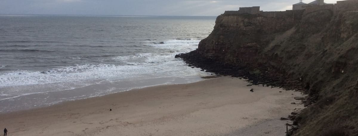 The quiet sands of King Edward’s Bay in Tynemouth on a calm winter day, with a single walker at the water’s edge beneath the headland cliff